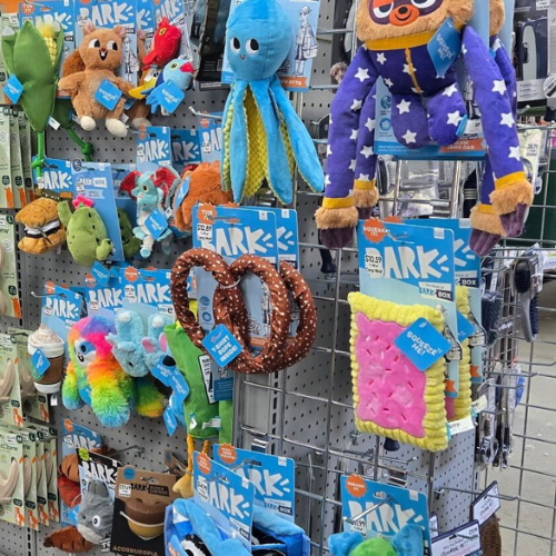 A colorful pet store aisle filled with various dog toys and accessories. Items include plush toys, chew toys, and balls, displayed on shelves and hooks.
