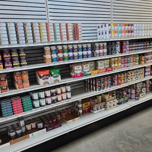 Store shelves filled with a variety of pet food cans and bags, neatly arranged by type and color. The setting is bright and orderly, conveying abundance.