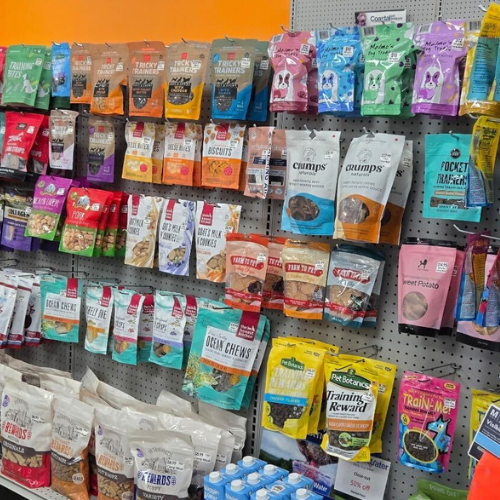 A display of various brightly packaged dog treats on a store shelf, featuring diverse flavors and brands against a vibrant orange wall, conveying abundance.