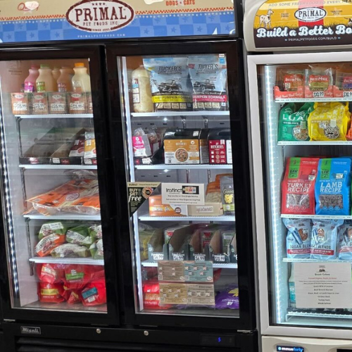 Refrigerated display case with various packaged pet foods, including raw meals, treats, and supplements, arranged neatly in bright, colorful packaging.