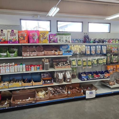 A well-stocked pet store aisle featuring shelves of colorful bags of pet food, treats, and toys. The setting appears organized and inviting.