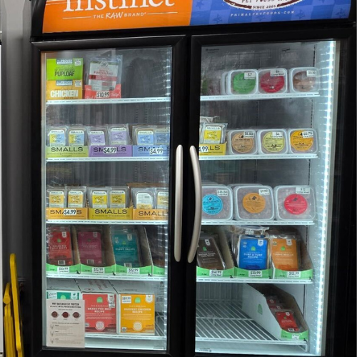 A well-stocked freezer with glass doors displays various pet food brands in uniform packaging. Brightly lit, sign reads 