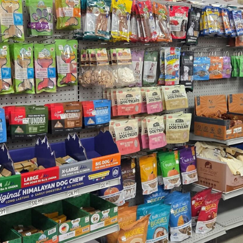 A well-stocked pet store display featuring a colorful variety of dog treats and chews. Packages in different shapes and sizes fill the shelves, conveying a sense of abundance and choice.