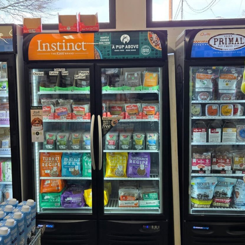 Refrigerated pet food section displaying brands like Instinct and Primal. Variety of colorful packaging with different flavors and options.