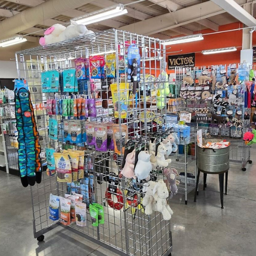 Pet store aisle displaying a variety of colorful pet toys, treats, and accessories on metal racks. The setting is bright and organized, conveying a welcoming atmosphere.